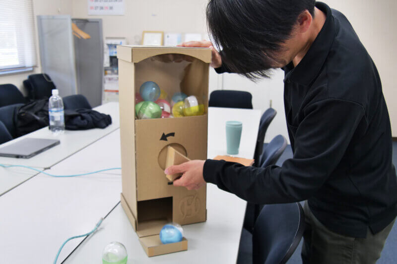 ダンボール製のガチャガチャを回す久保
